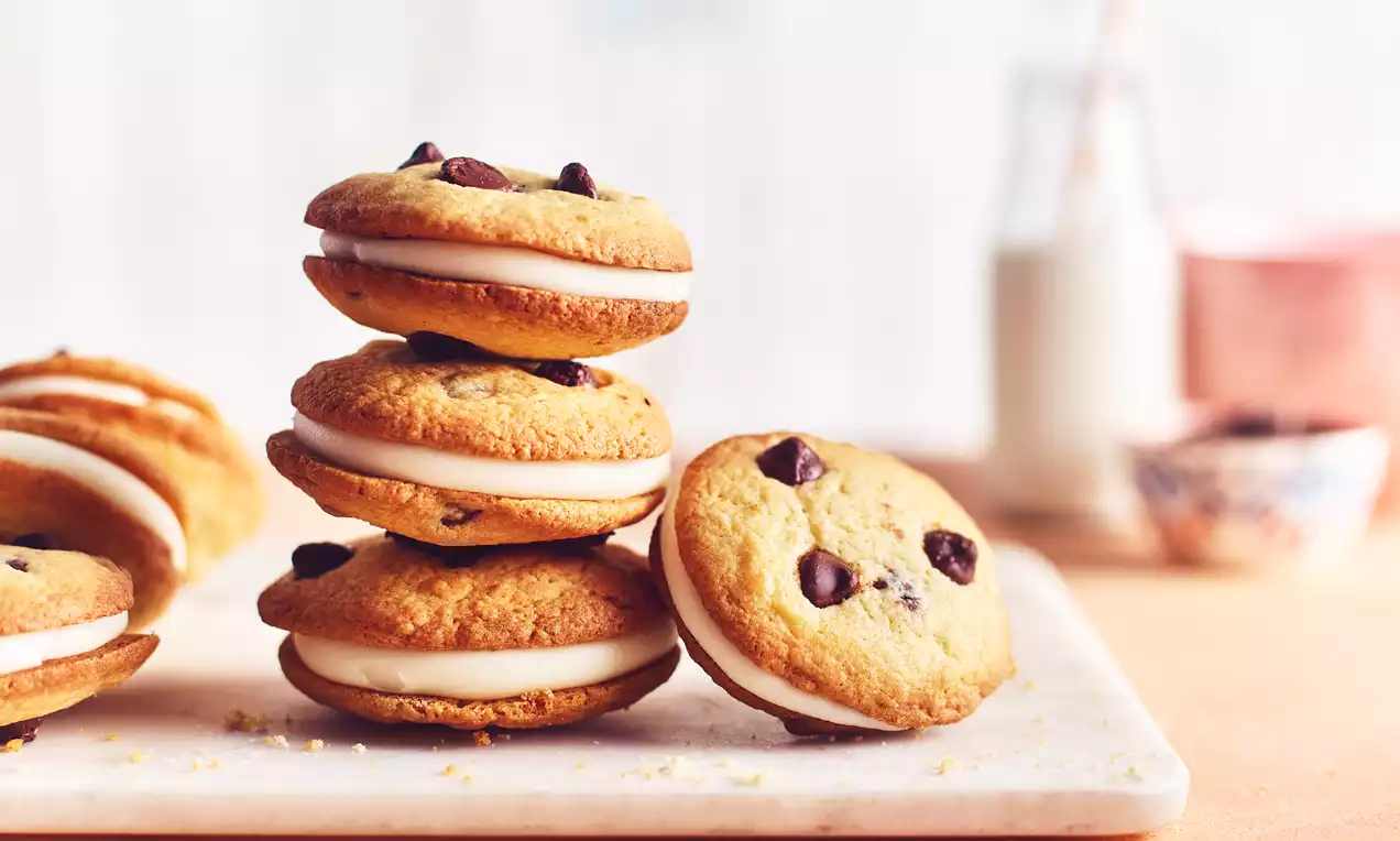 Golden chocolate chip cookie sandwiches stacked and arranged on a white marble board, filled with smooth white cream filling