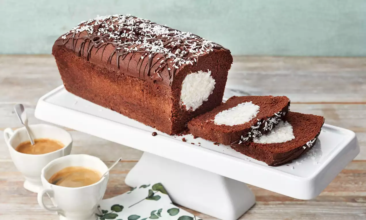 Rechteckiger Schoko-Kokos-Kuchen mit saftiger, dunkler Kruste, heller Kokosfüllung und Schokoglasur auf weißer Kuchenplatte angerichtet