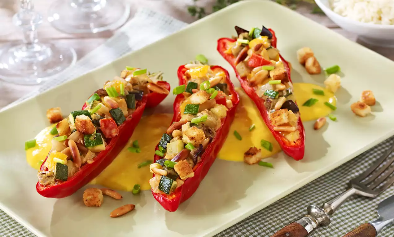 Drei rot glänzende, halbierte Paprikaschoten mit bunt gebratenem Gemüse und goldbraunen Croutons auf heller Platte mit Kräuterdekor