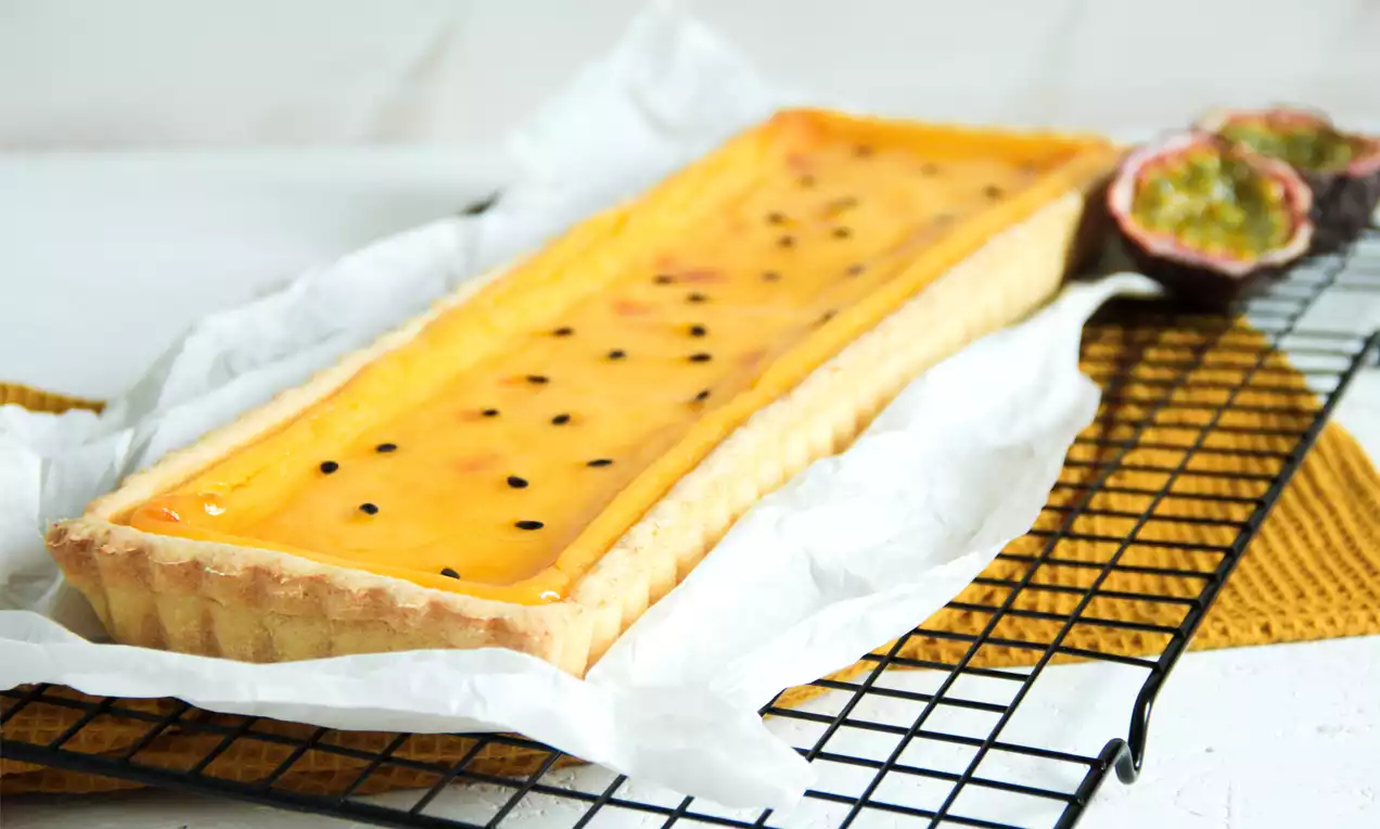 Rechteckige Passionsfruchttarte mit goldgelber Creme und schwarzen Kernen in Mürbeteigkruste auf Backpapier und Gitterrost drapiert
