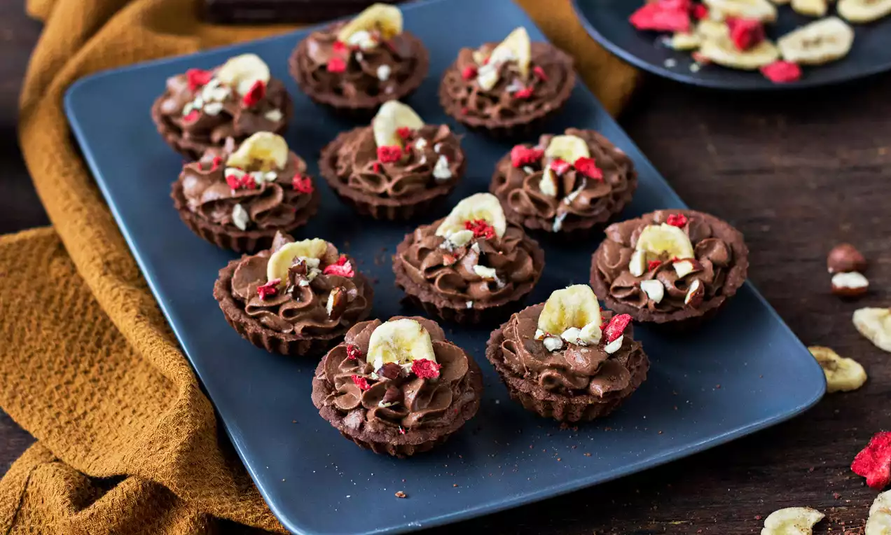 Fit kakaové košíčky s tmavohnedým krémom zdobené plátkami banánu, červenými kúskami ovocia a orieškami, usporiadané na tmavomodrom tanieri