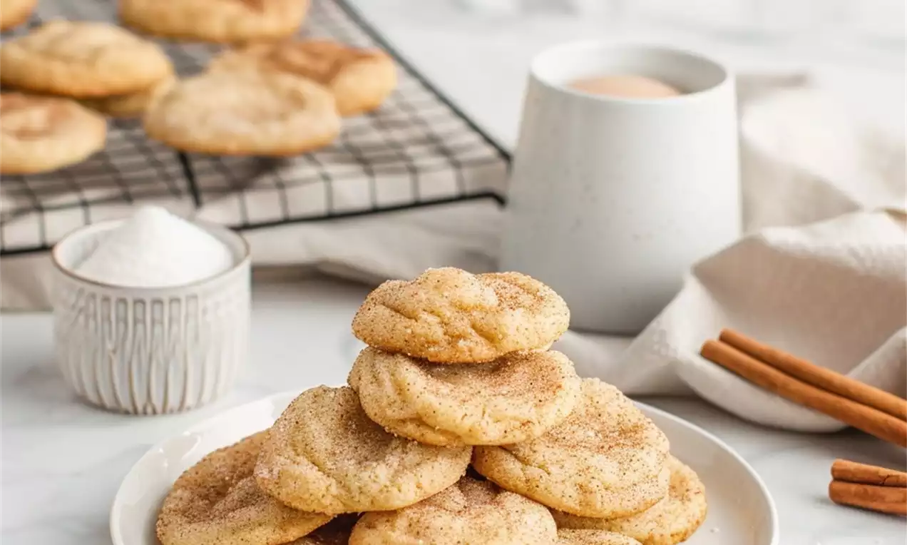 Golden brown snickerdoodles with crinkled tops stacked on a white plate, surrounded by cinnamon sticks, sugar, and a mug on a marble surface