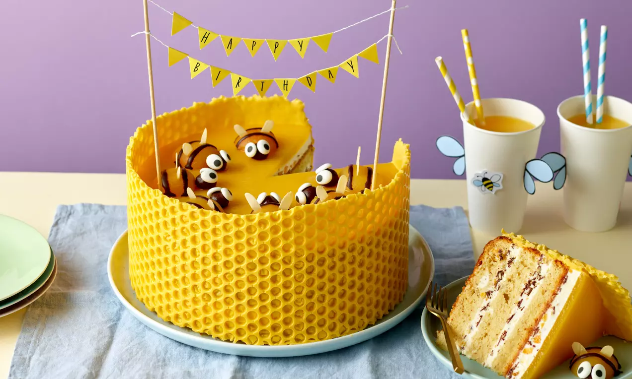 Runde Bienen-Torte mit leuchtend gelber Wabenverzierung, dekoriert mit kleinen Bienenfiguren und Wimpeln auf hellblauer Serviette angerichtet
