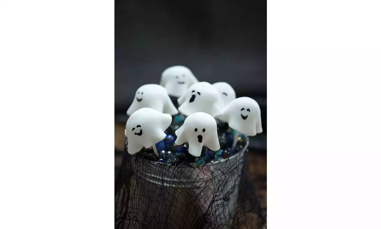 White ghost-shaped cake pops with smooth, flowing forms, drawn faces, arranged on sticks in a dark metallic container draped in netting