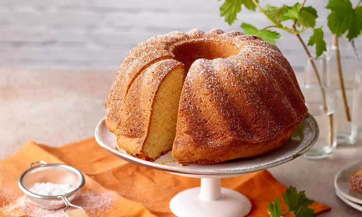 Goldbrauner Gugelhupf mit zarter Puderzuckerhaube auf weißer Tortenplatte, angeschnitten und von frischen grünen Blättern umgeben