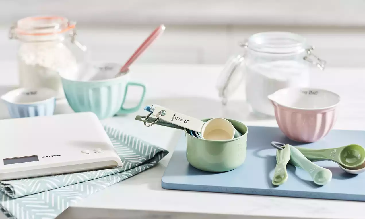 Pastel measuring utensils arranged on a blue board with flour jars, a white digital scale, and a patterned cloth on a white surface