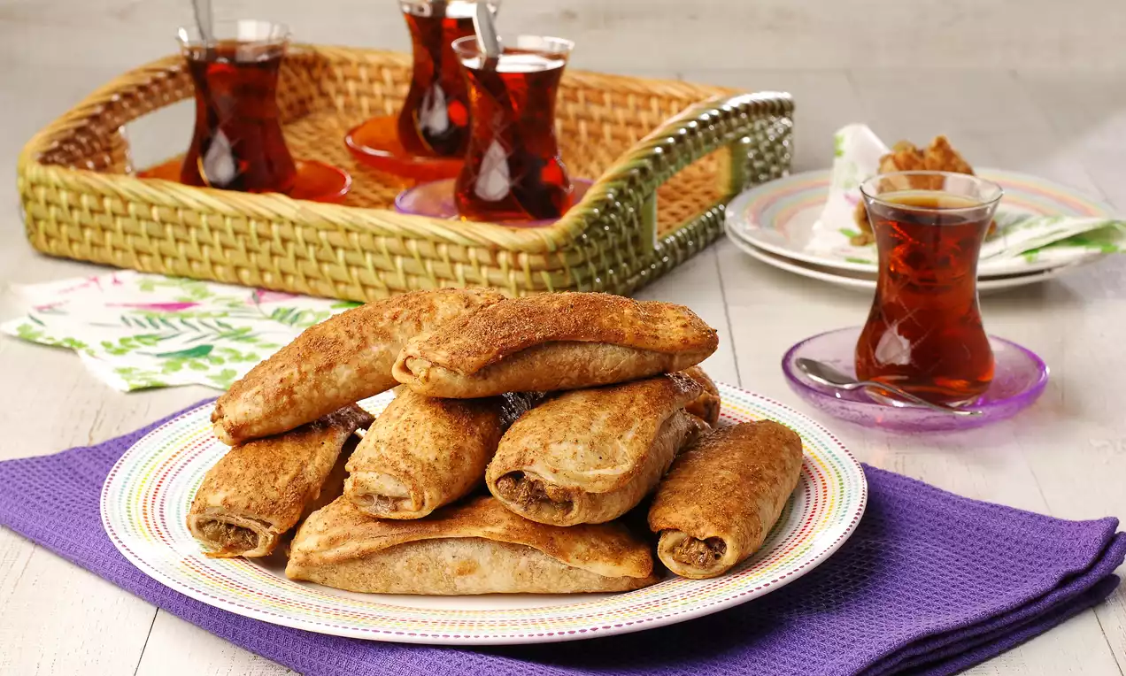 Altın kahverengi mercimekli börekler, çizgili bir tabakta, mor bir örtü üzerinde sunulmuş, yanında ince belli çay bardaklarıyla birlikte