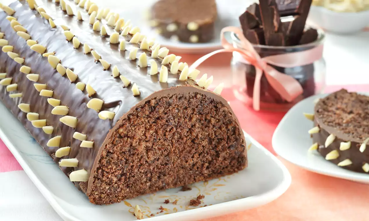 Czekoladowy rulon o półokrągłym kształcie z polewą czekoladową i dekoracją z jasnych migdałów na białym prostokątnym talerzu