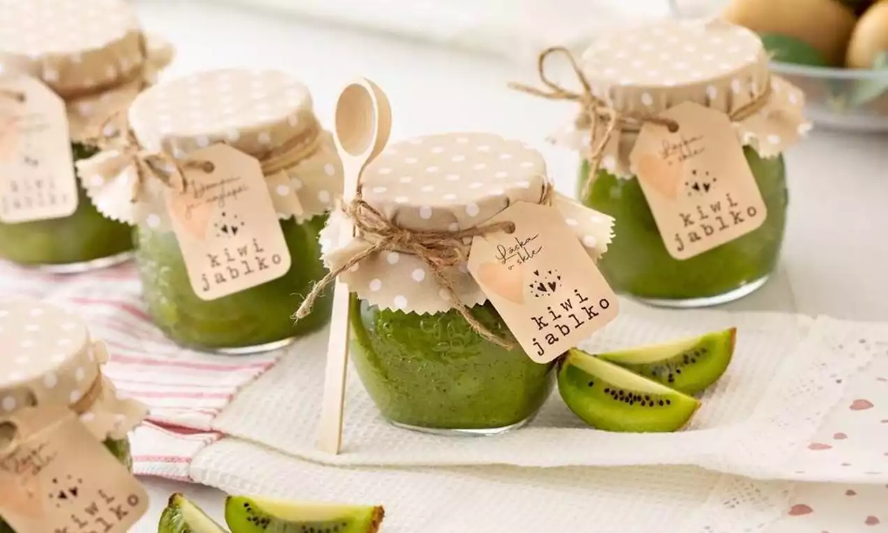 Sklenené poháre s jasne zeleným kiwi-jablkovým džemom, ozdobené bodkovanými béžovými viečkami a kartičkami, položené na bielej utierke s plátkami kiwi