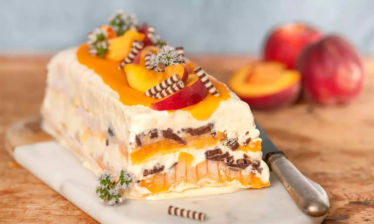 Vanille-Parfait mit heller Cremeschicht, Schokoladenstücken, frischen Pfirsichschnitzen und Blüten auf einem weißen Servierbrett