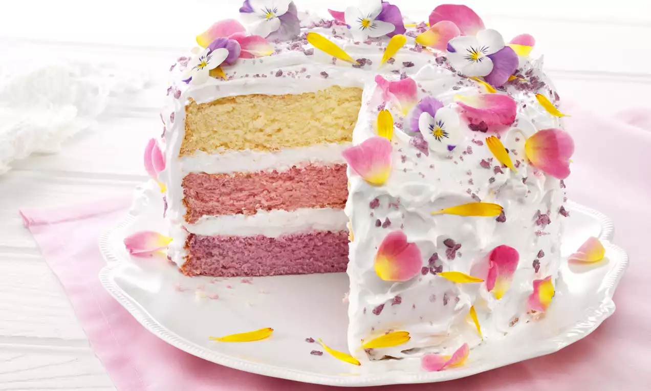 Troslojna torta na bijelom tanjiru, prekrivena šlagom i ukrašena šarenim jestivim cvijećem, s toplim bojama roze i žute u presjeku