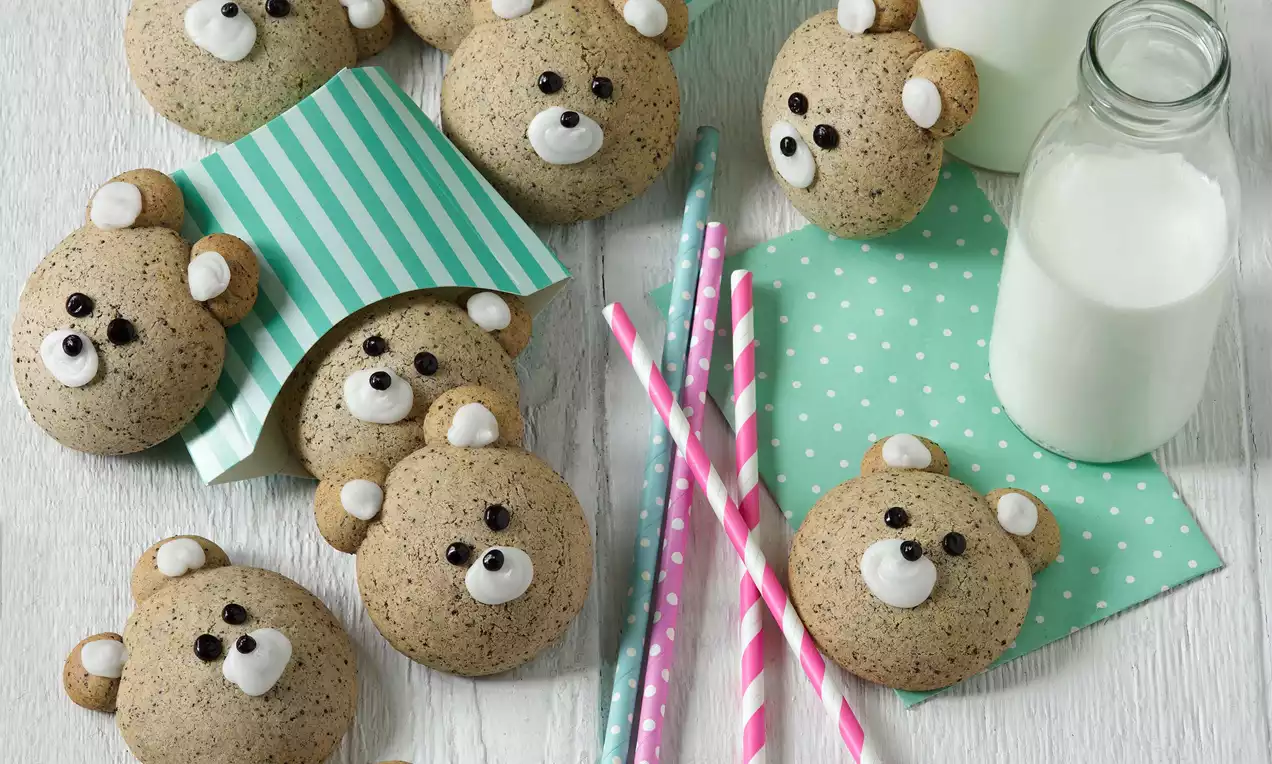 Makové perníčky vo svetlohnedom odtieni, tvarované ako medvedie hlavičky s čiernymi a bielymi detailmi na očiach a ňufákoch, položené na svetlom drevenom povrchu vedľa farebných slamiek, servítok a fľaší s mliekom