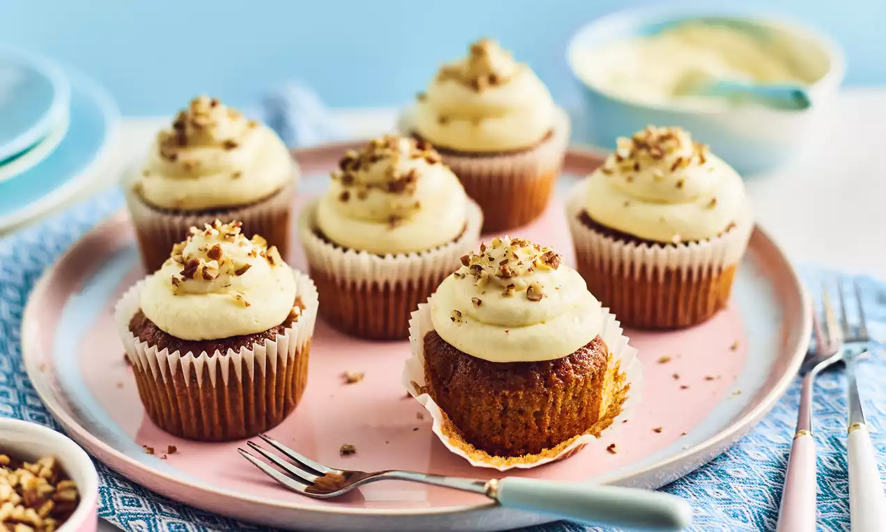Carrot cake cupcakes with golden brown bases, swirled cream cheese frosting, and chopped nuts on a pink plate with a fork nearby