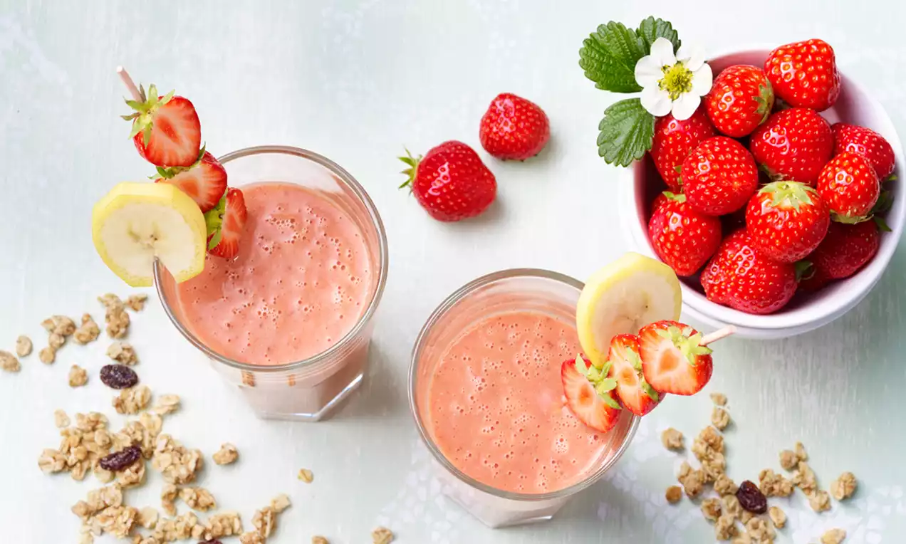 Cam bardaklarda açık pembe çilekli smoothie, üzerine çilek ve muz dilimli şişler yerleştirilmiş, yanında kasede taze çilekler ve granola bulunan düzenleme