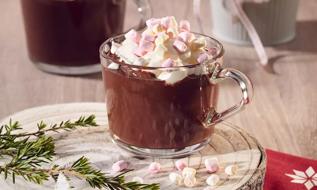 Átlátszó üvegbögrében gőzölgő, sötétbarna forró csokoládé selymes tejszínhabbal és rózsaszínes-fehér pillecukorral fenyőággal egy fatányéron