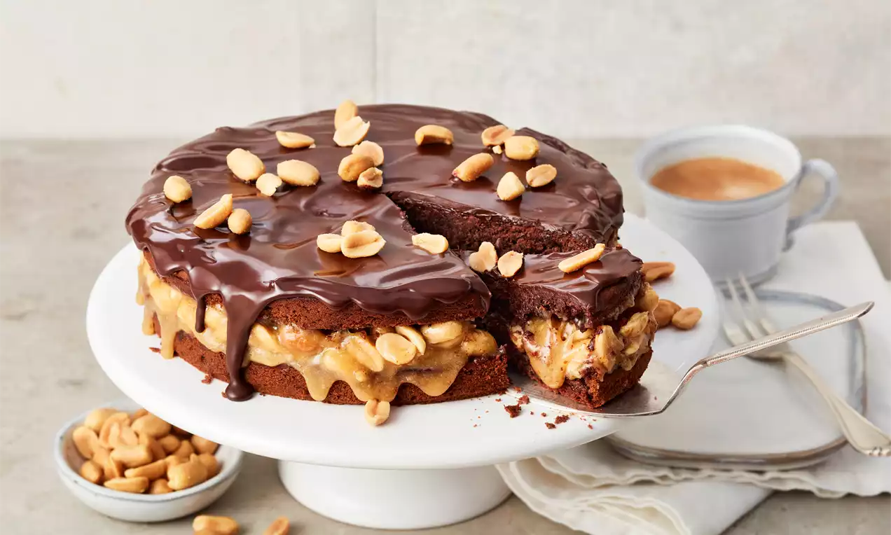 Runder Schokoladenkuchen mit glänzender Glasur, gefüllt mit cremiger Erdnussmasse, garniert mit ganzen Erdnüssen auf weißer Kuchenplatte