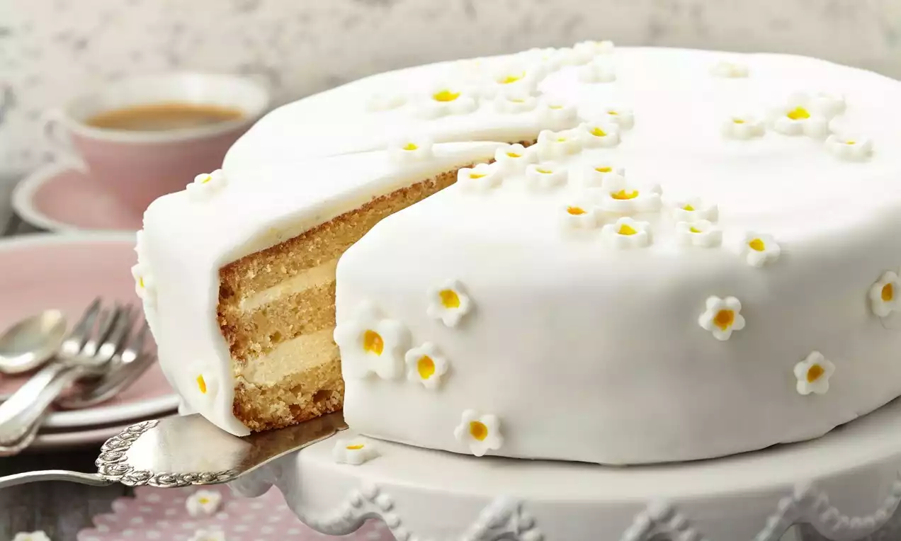 Kulatý dort s bílým fondánovým povrchem, zdobený drobnými květy s žlutým středem, na zdobeném stojanu vedle růžového šálku kávy