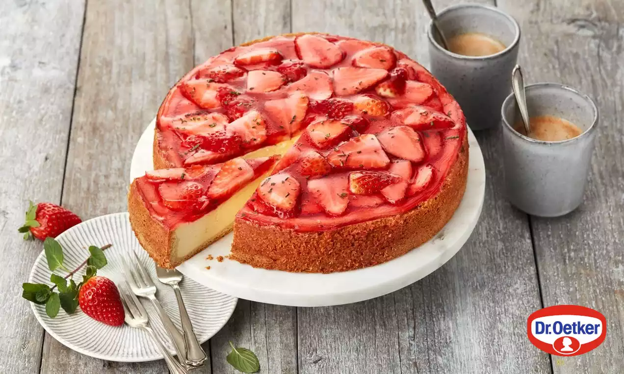 Cheesecake s krémově světlou náplní na hnědém sušenkovém základu, pokrytý lesklou červenou jahodovou polevou a plátky jahod na bílém podnosu