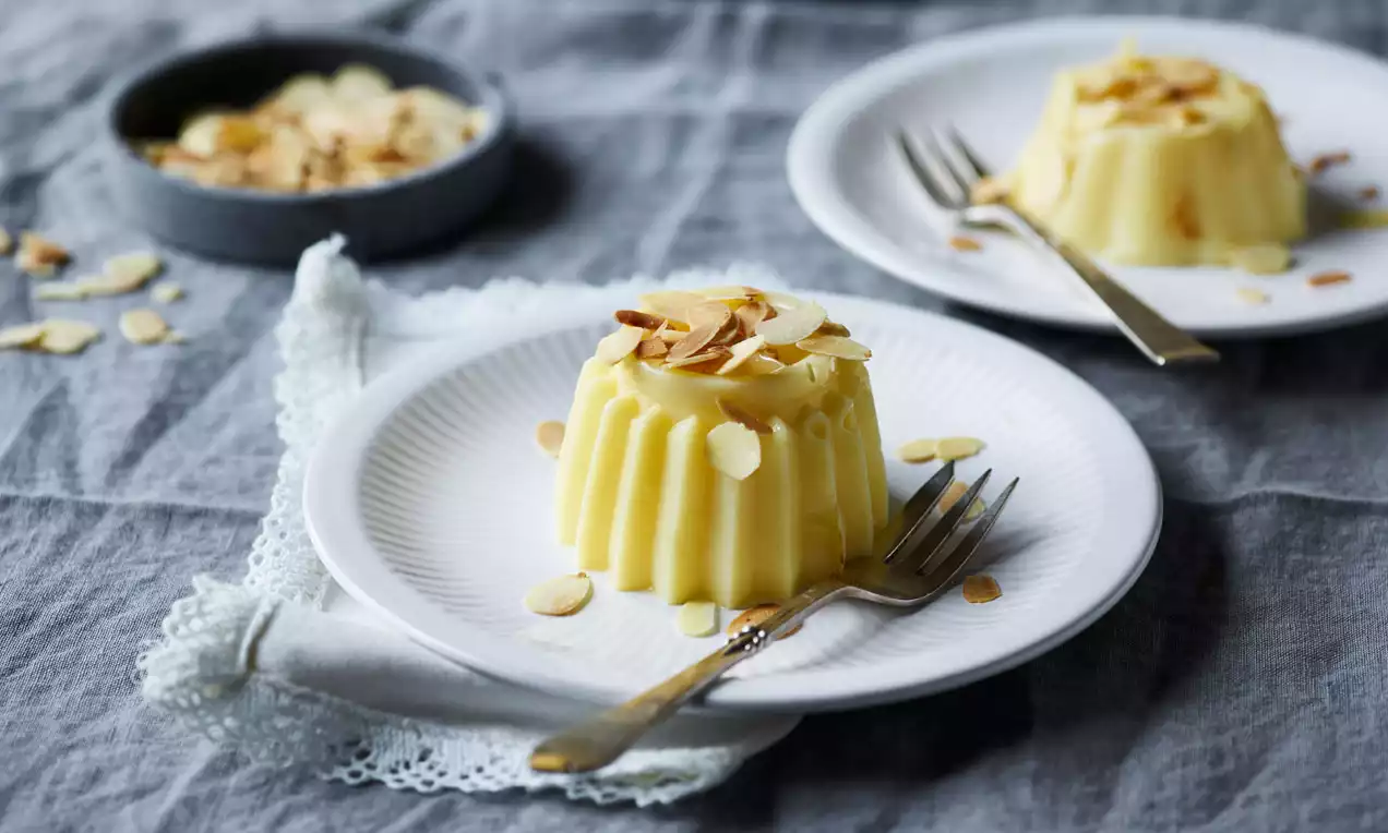 Budino cremoso giallo chiaro con superficie liscia e bordi ondulati, decorato con sottili scaglie di mandorla su un piatto bianco con forchetta dorata