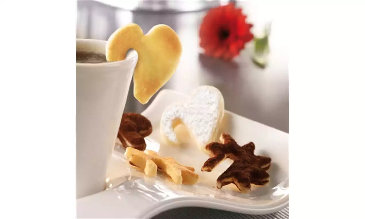 Herzförmiger Keks am Tassenrand, daneben helle und dunkle Kekse in Blattform auf weißem Teller, im Hintergrund rote Blume