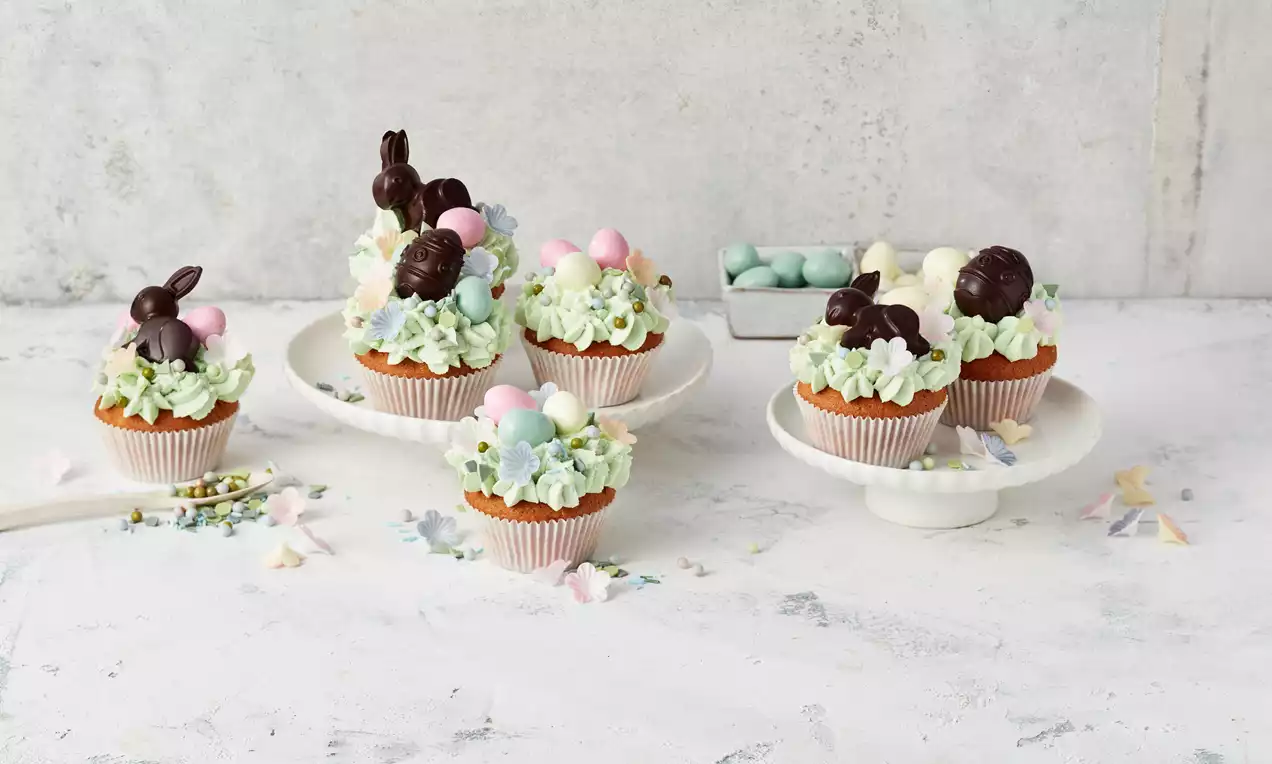 Muffini zlatnosmeđe boje u papirnatim košaricama, ukrašeni svijetlozelenim kremom, čokoladnim figurama i pastelno obojenim jajima