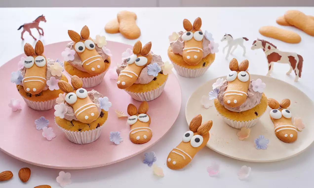 Süße Muffins mit Pferdegesichtern aus hellen Keksen, Mandeln und Zuckerdeko auf rosa und weißem Teller mit Blütenstreudekoration