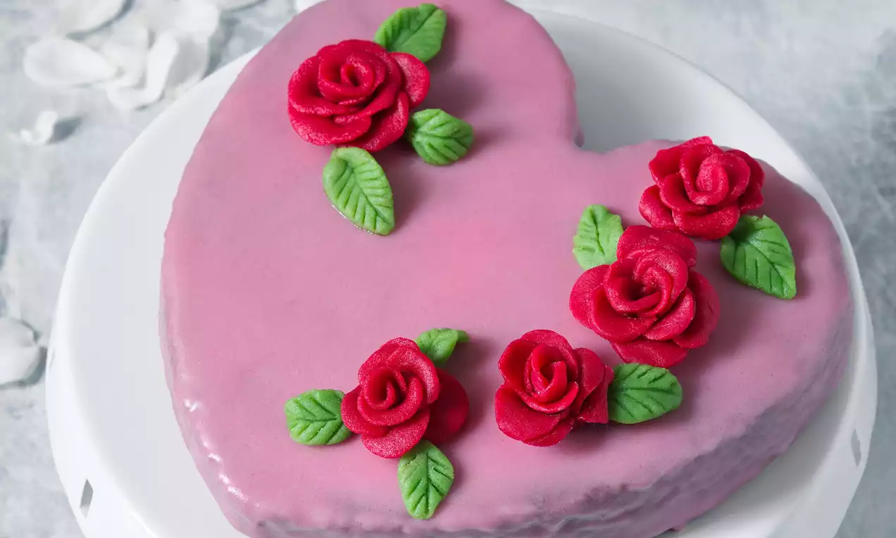 Koláč v tvare srdca s hladkou fialovoružovou polevou, ozdobený červenými ružami a zelenými lístkami na bielom podklade