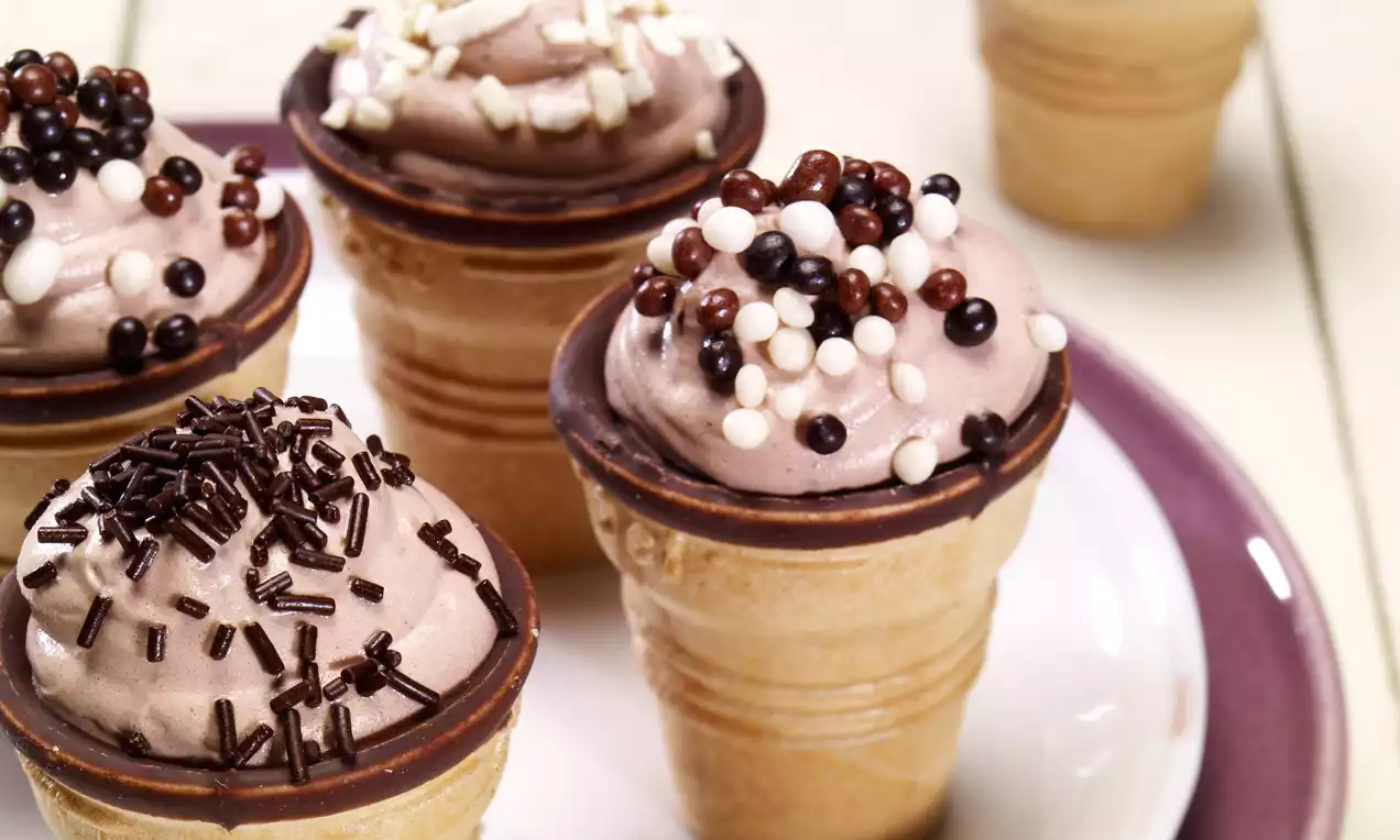 Schokocreme in zartbraunen Waffelbechern mit Schokoladenrand, garniert mit bunten Schokokugeln und Schokostreuseln auf weißem Teller