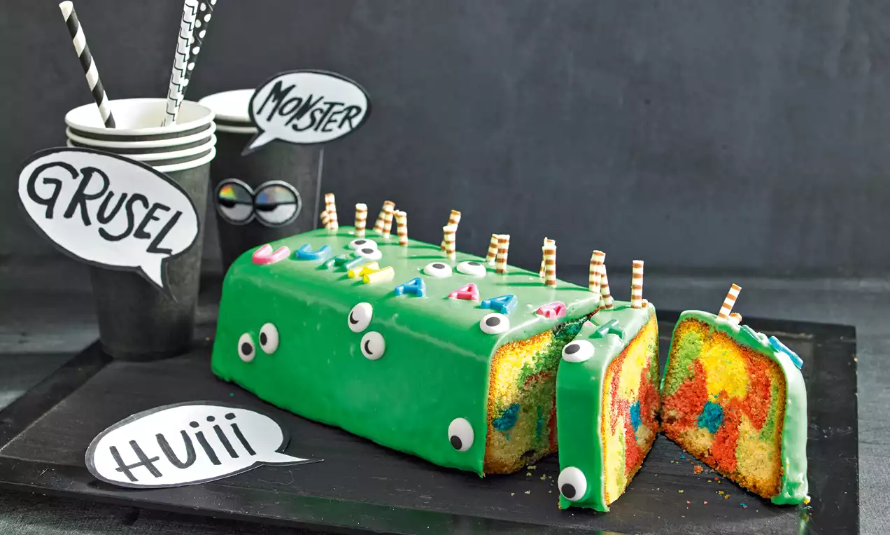 Rechteckiger Kuchen mit grüner Glasur, buntem Innenleben, dekoriert mit Zuckeraugen und Schokoröllchen auf schwarzer Platte