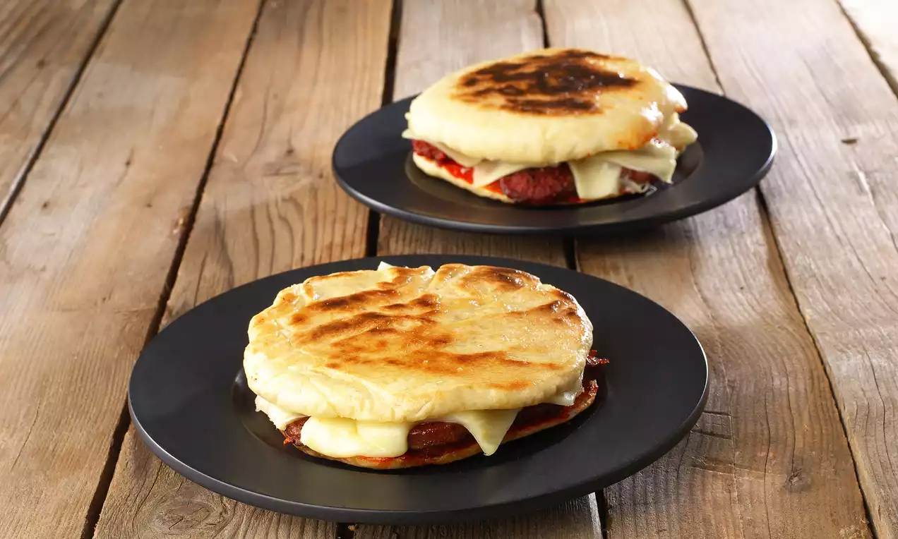 Altın renginde kızarmış bazlama arasında eritilmiş peynir ve sucuk dilimleri, siyah tabaklarda rustik ahşap zeminde sunulmuş