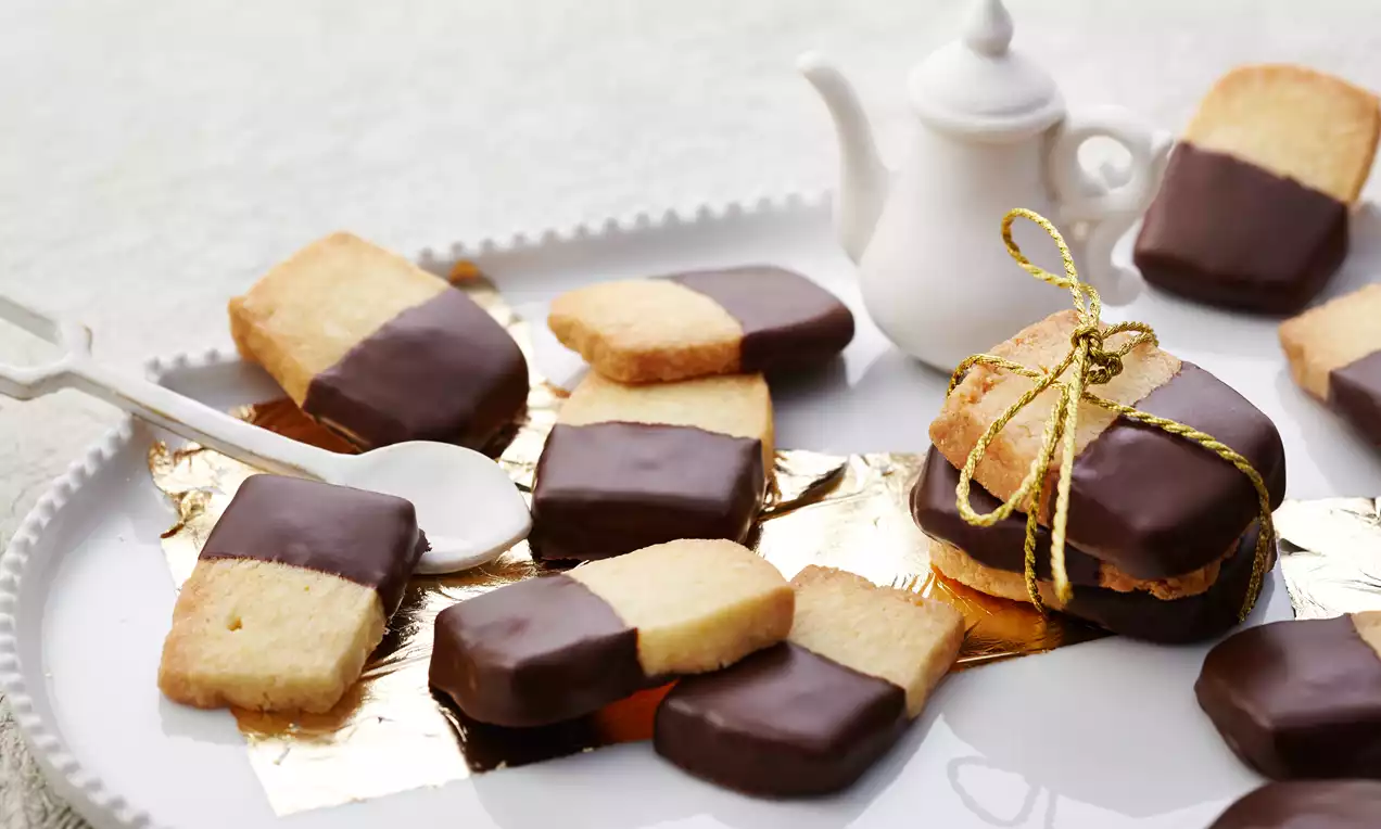 Feines Mandelgebäck in rechteckiger Form, halb mit glänzender dunkler Schokolade überzogen, dekorativ auf weißem Teller mit goldener Kordel arrangiert