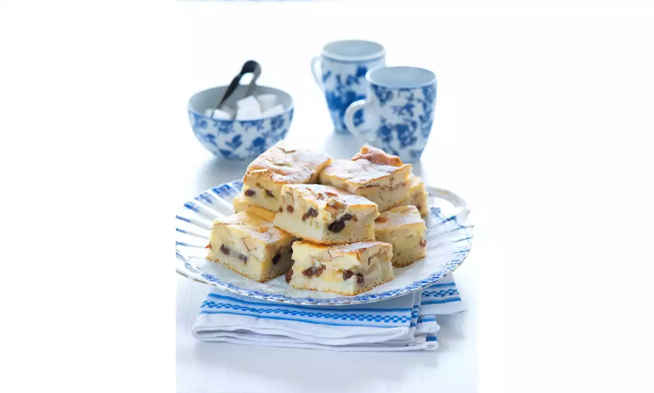 Štvorce zlatistej bublaniny s hrozienkami uložené na vzorovanom sklenenom tanieri, na bielo-modrom obrúsku vedľa porcelánových šálok