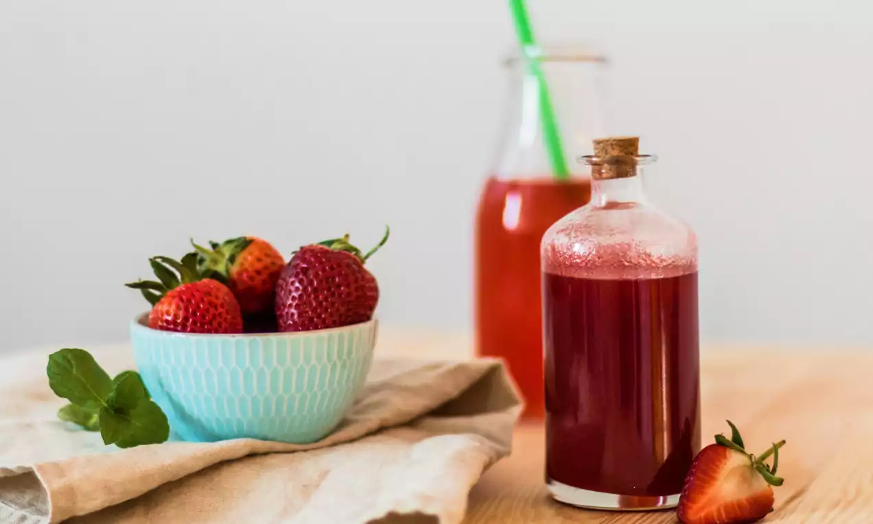 Matně červený jahodový sirup v malé skleněné lahvi s korkovým uzávěrem vedle čerstvých jahod ve světle modré misce na béžové látce