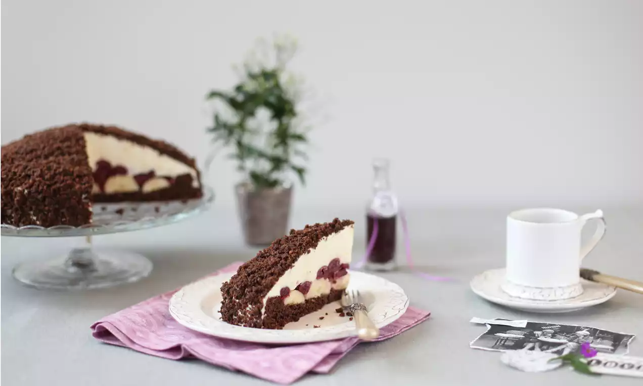 Ein Stück Maulwurfkuchen mit heller Bananencreme, roten Sauerkirschen und dunklen Schokobröseln auf einem weißen Teller mit rosa Serviette
