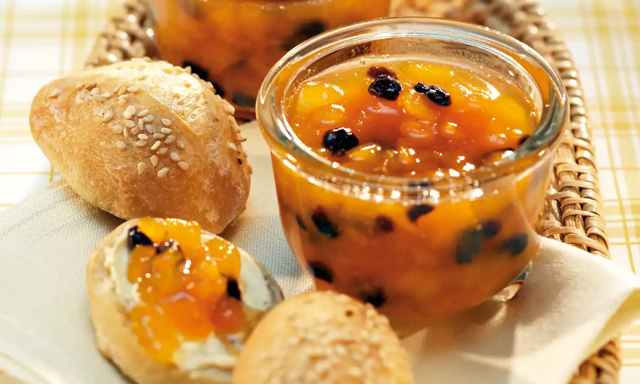 Marmellata dorata di albicocche con pezzetti lucidi, arricchita da uvetta scura e cannella, servita in vasetti di vetro accanto a panini al sesamo