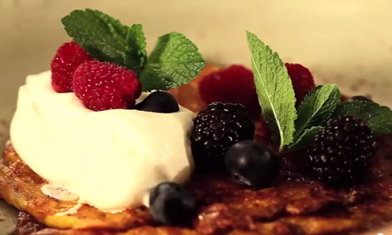 Gyllene saffranspannkaka toppad med vit grädde, röda hallon, svarta björnbär, blåbär och gröna myntablad på en ljus tallrik