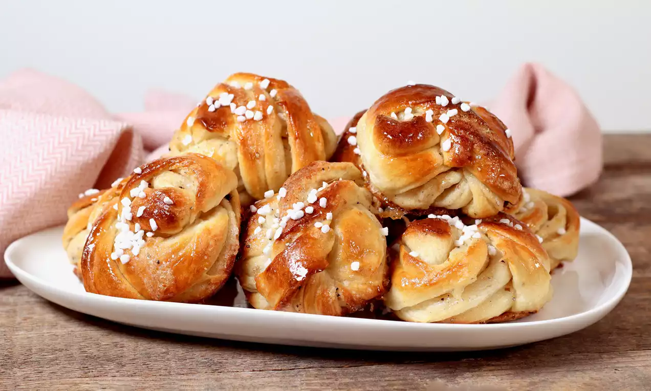 Kierretyt kardemummapullat kullanruskeita ja kiiltäviä, myötäilevät valkoisen lautasen muotoa, pinnalla rosoista raesokeria.