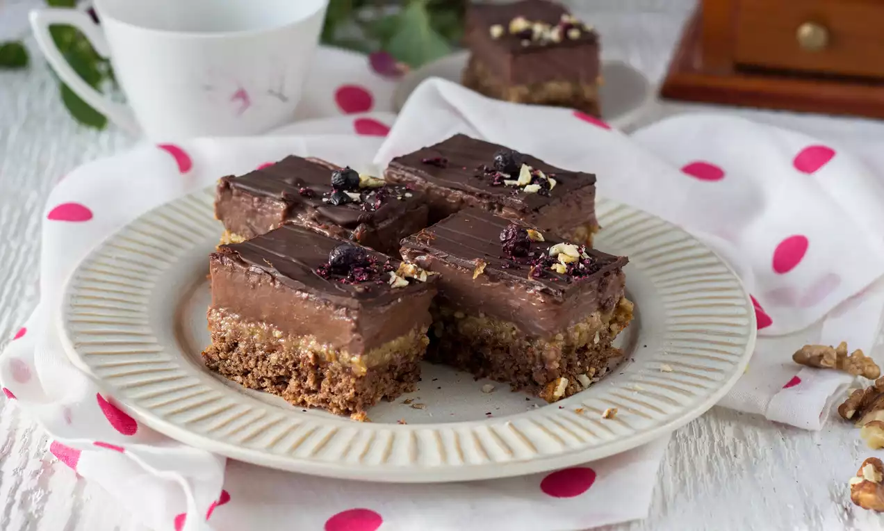 Hnedasté orechové kocky s lesklou tmavou čokoládovou vrstvou, posypané krájanými orechmi a sušenými brusnicami na bledom porcelánovom tanieri