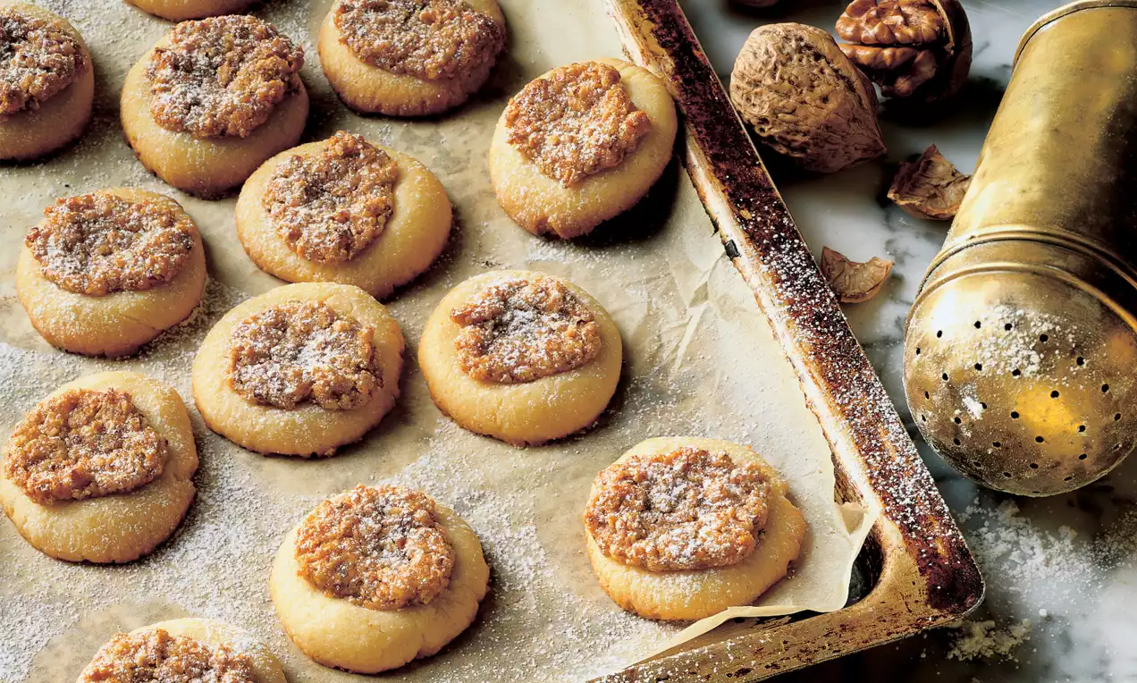 Runde Walnussplätzchen mit goldbrauner Füllung auf leicht gebräuntem Backpapier, umgeben von zerbrochenen Walnüssen und einem Metallstreuer