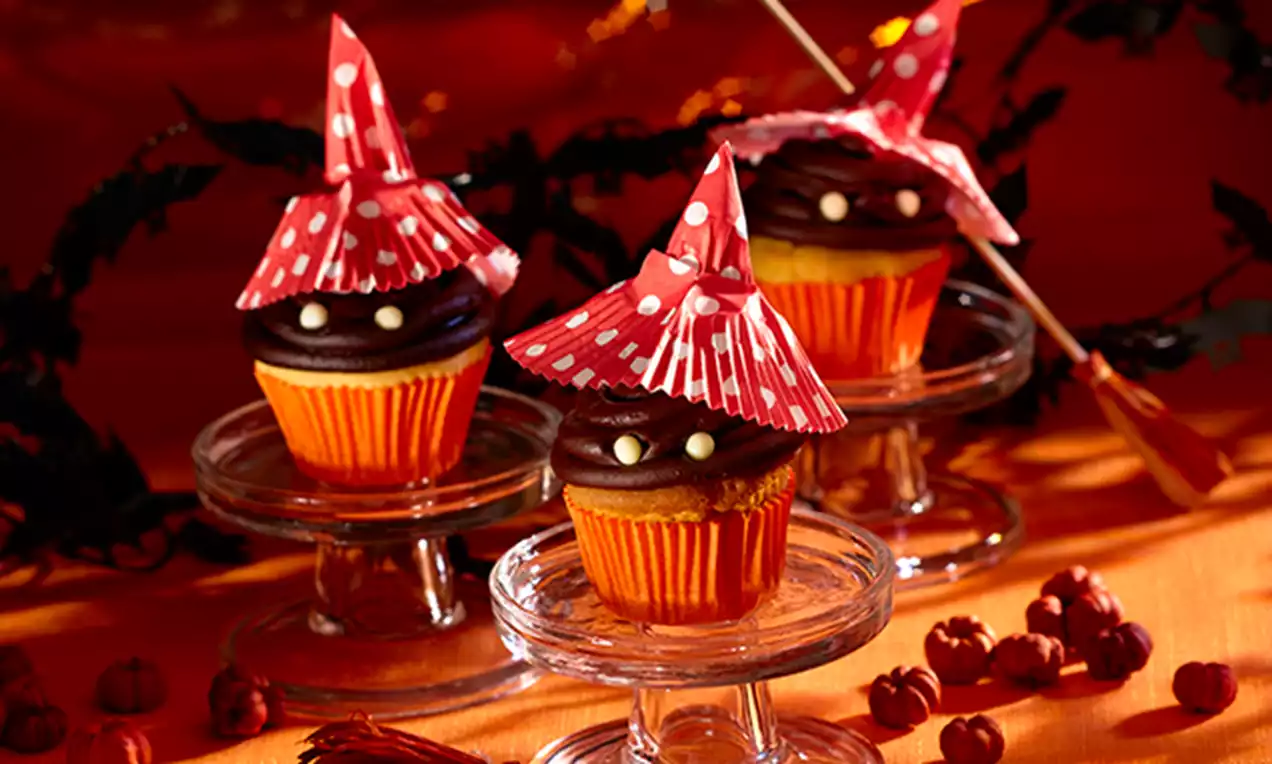 Cupcakes orange au chocolat surmontés de chapeaux rouges à pois, posés sur des présentoirs transparents parmi des mini citrouilles