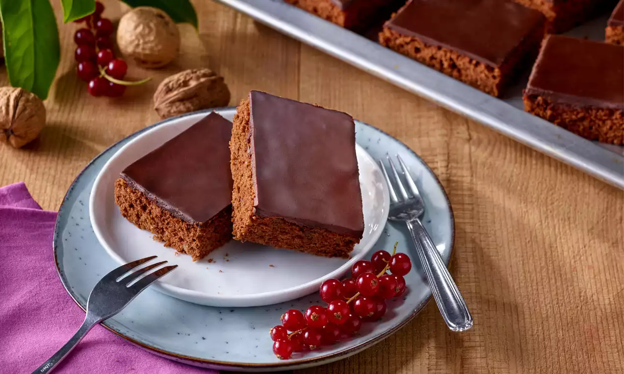Zwei rechteckige Stücke dunkler Schokokuchen mit glänzendem Guss auf einem Teller mit roten Johannisbeeren und Silberbesteck angerichtet