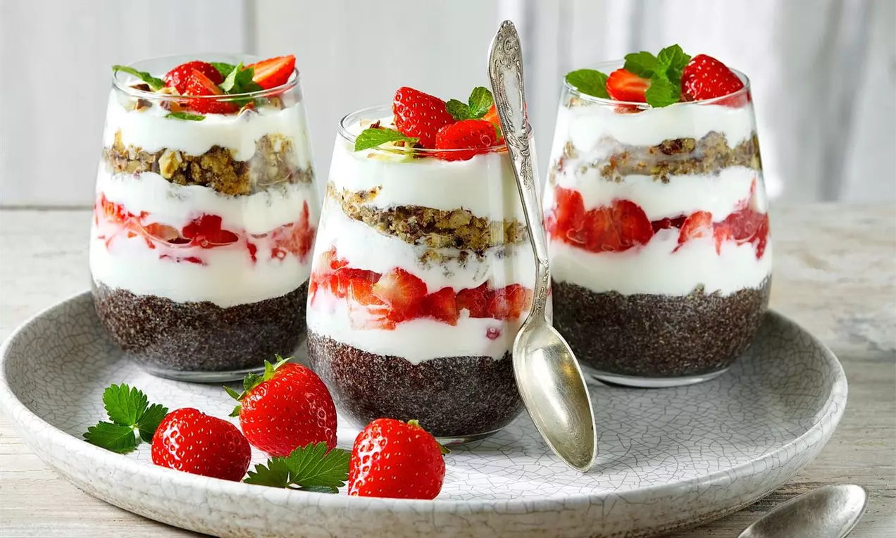 Jemné vrstvy bieleho jogurtu, červených jahôd a chrumkavého posýpkového základu v sklených pohároch na šedom keramickom tanieri