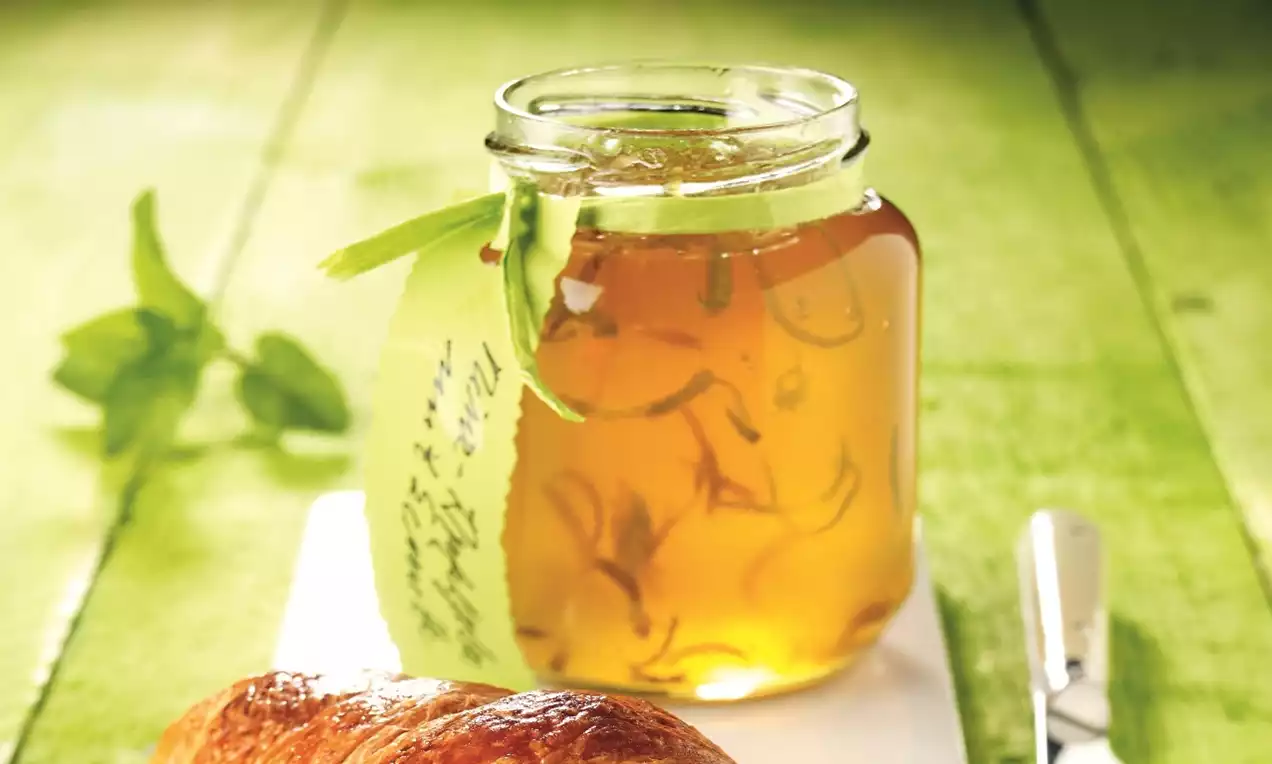 Jeleu auriu translucid într-un borcan de sticlă, presărat cu bucăți fine, decorat cu mentă proaspătă și etichetat pe fundal verde