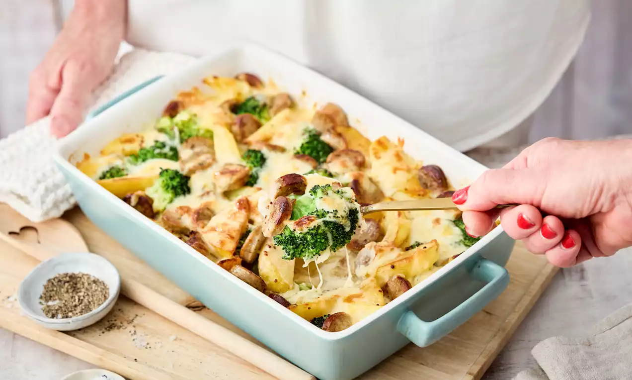 Bratwurst-Auflauf mit goldbraun überbackenem Käse, grünen Brokkoliröschen und Kartoffelspalten in einer rechteckigen hellblauen Form