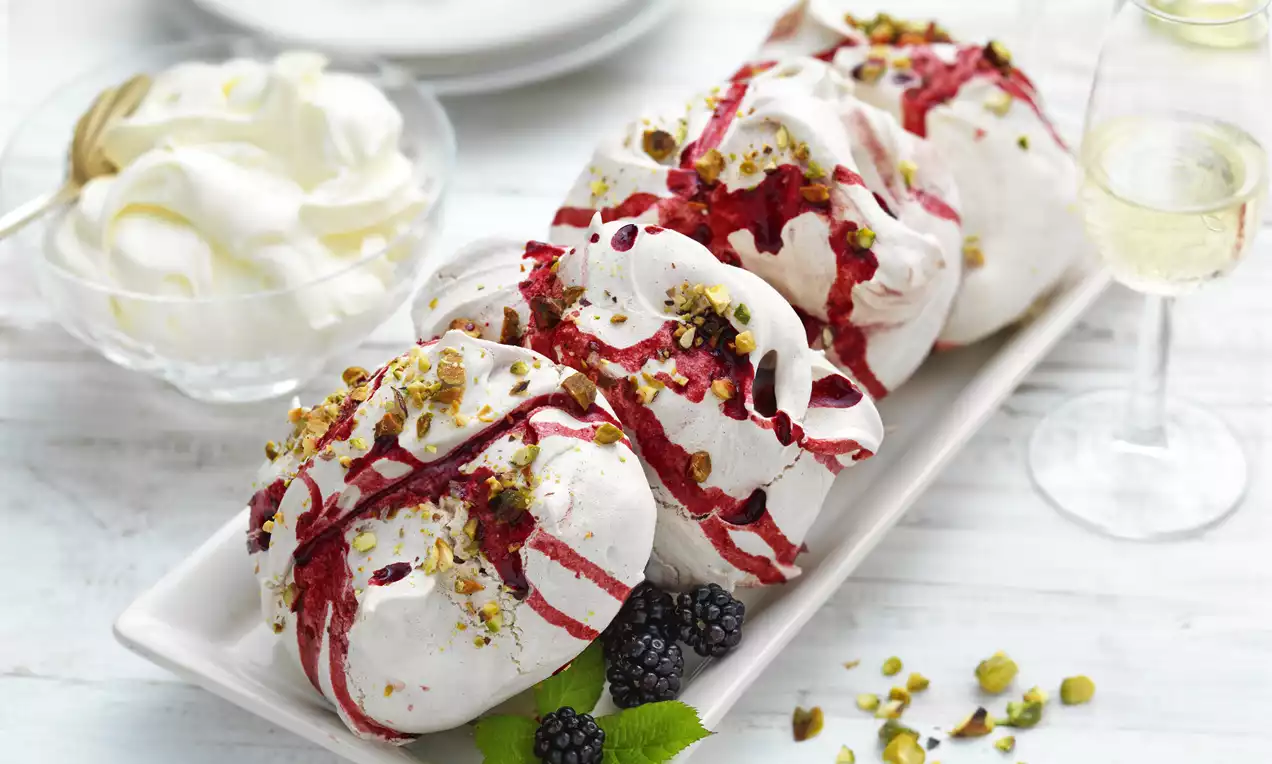 White meringues swirled with deep red blackberry streaks, topped with chopped pistachios, arranged on a rectangular white plate