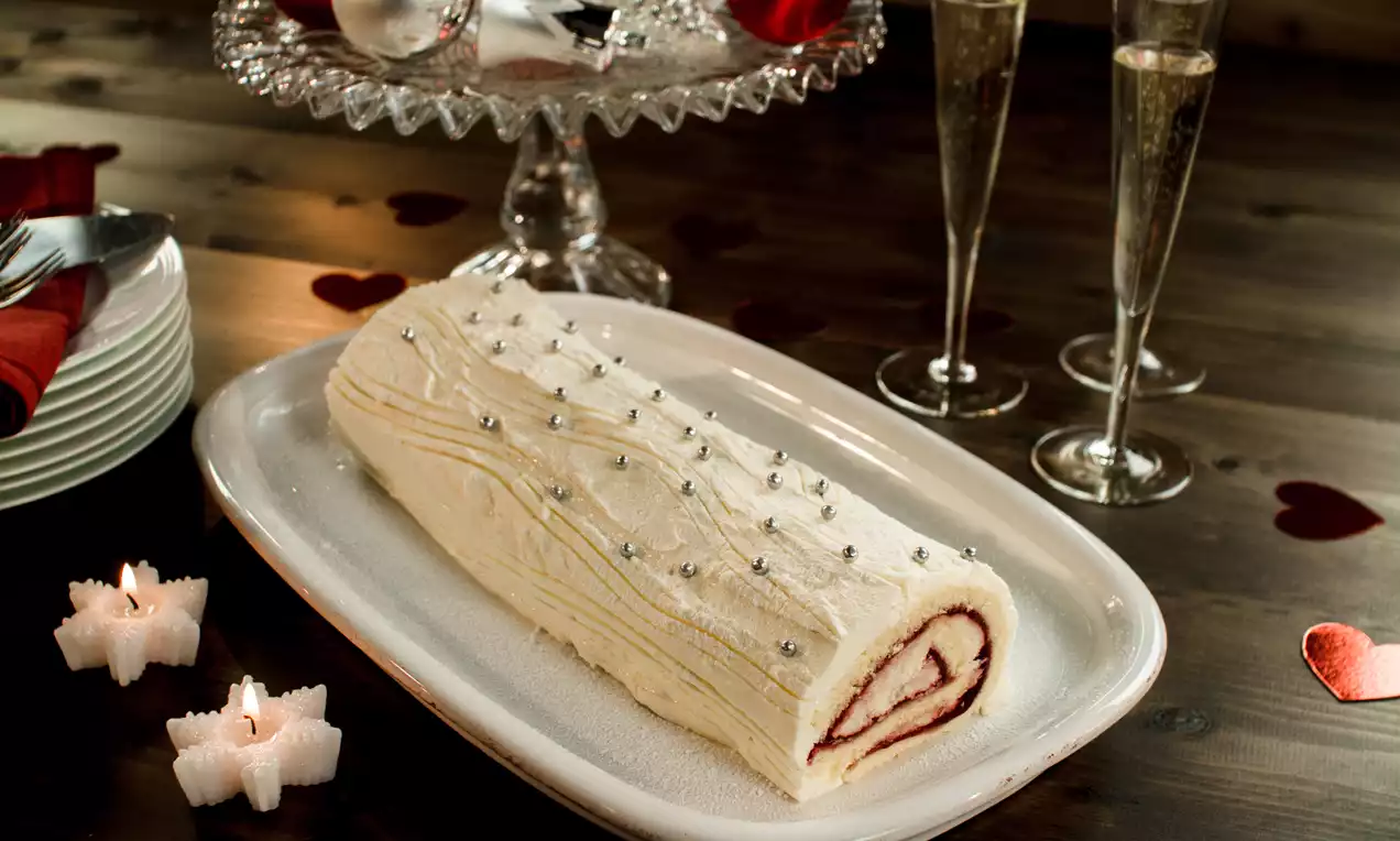 Kerststronk met witte botercrème-laag, licht golvend en bestrooid met zilveren parels, op een groot wit bord met feestelijke decoratie
