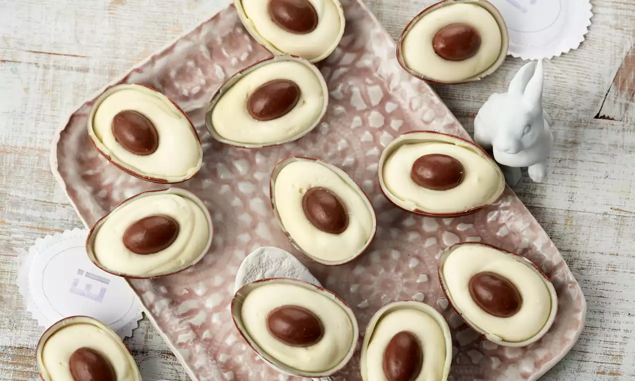 Halbe Schoko-Eierschalen gefüllt mit heller Creme und dekoriert mit einem glatten braunen Schokoladenei auf rosa gemusterter Platte