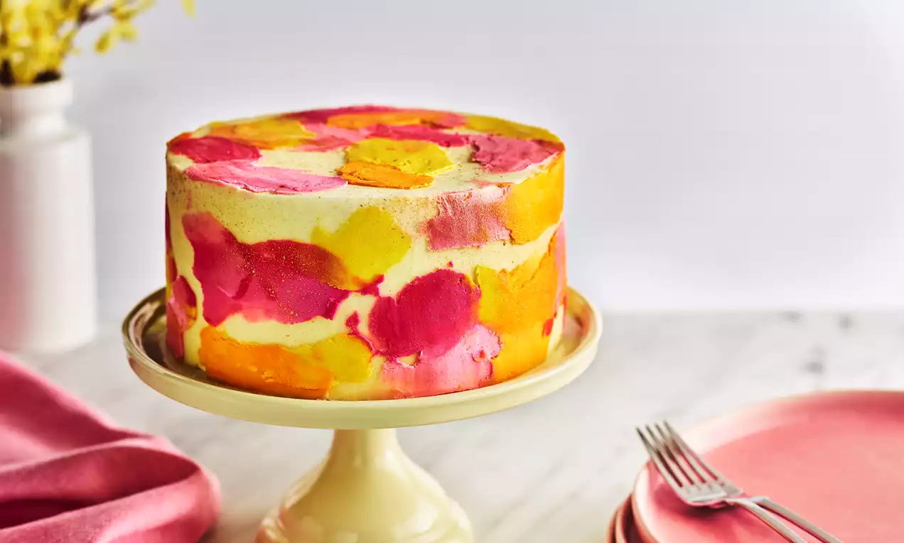 Round cake with a smooth buttercream surface painted in overlapping bold pink, orange, and yellow strokes on a cream cake stand
