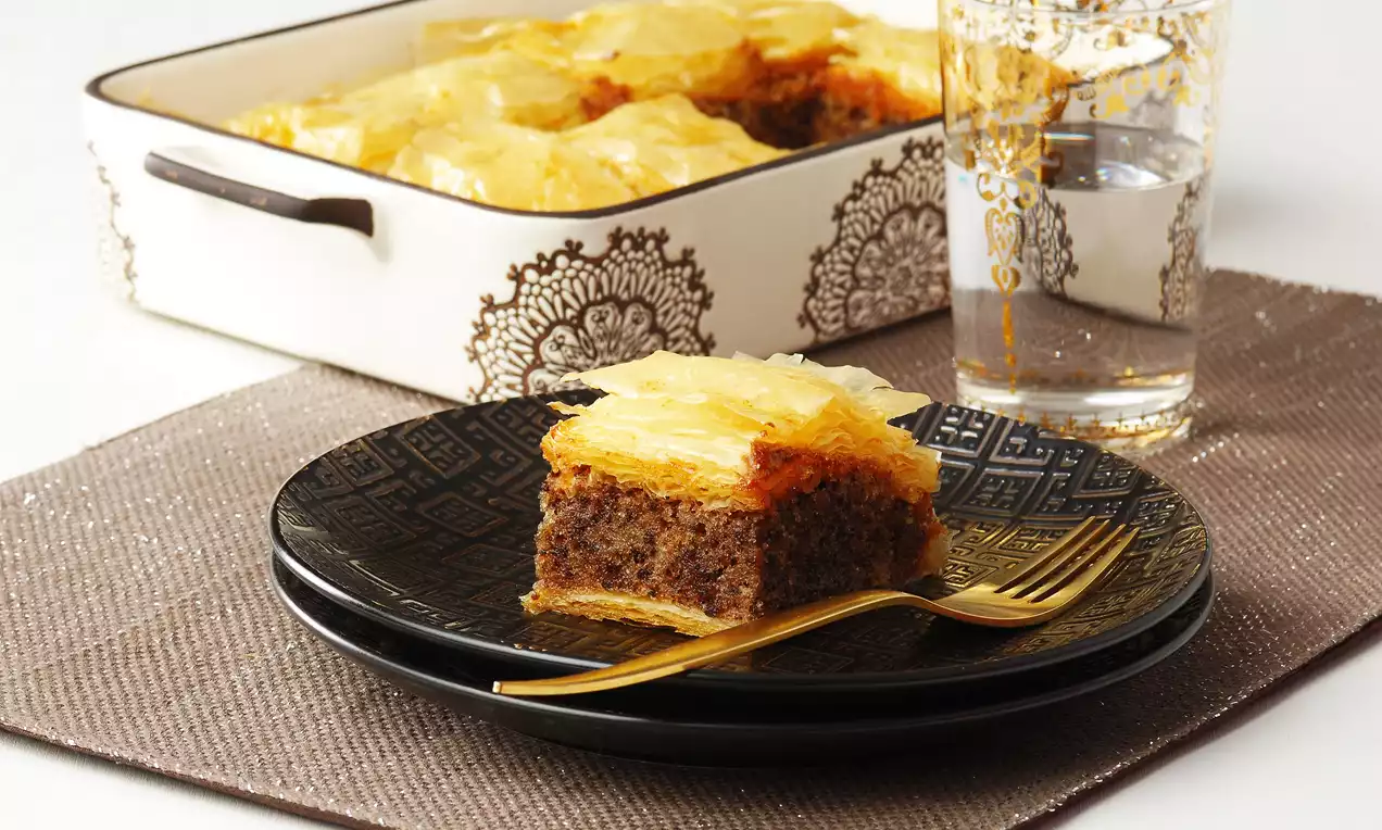 Kekli baklava, siyah desenli tabakta altın renkli çatalla, şeffaf desenli bir bardak su eşliğinde sunulmuş, üstü parlak ve kıtır katmanlı