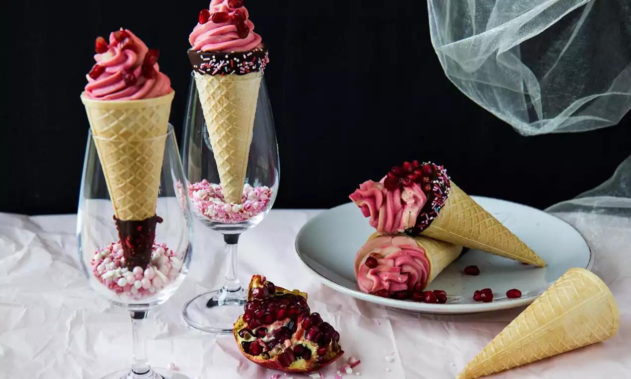 Kornouty ze světlého vaflového těsta s náplní růžového krému ozdobené granátovým jablkem, servírované ve sklenicích a na talíři s dekorací